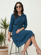 Woman wearing a blue dress sitting on a stool with a plant in the background
