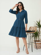 Woman wearing a blue dress standing in a minimalistic room with a plant and chair.