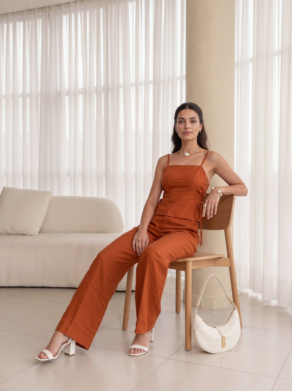 A woman models a rust-orange sleeveless jumpsuit while sitting on a wooden chair in a bright, modern room with white sheer curtains and a cream sofa. She is wearing white heeled sandals and a white handbag rests on the chair.