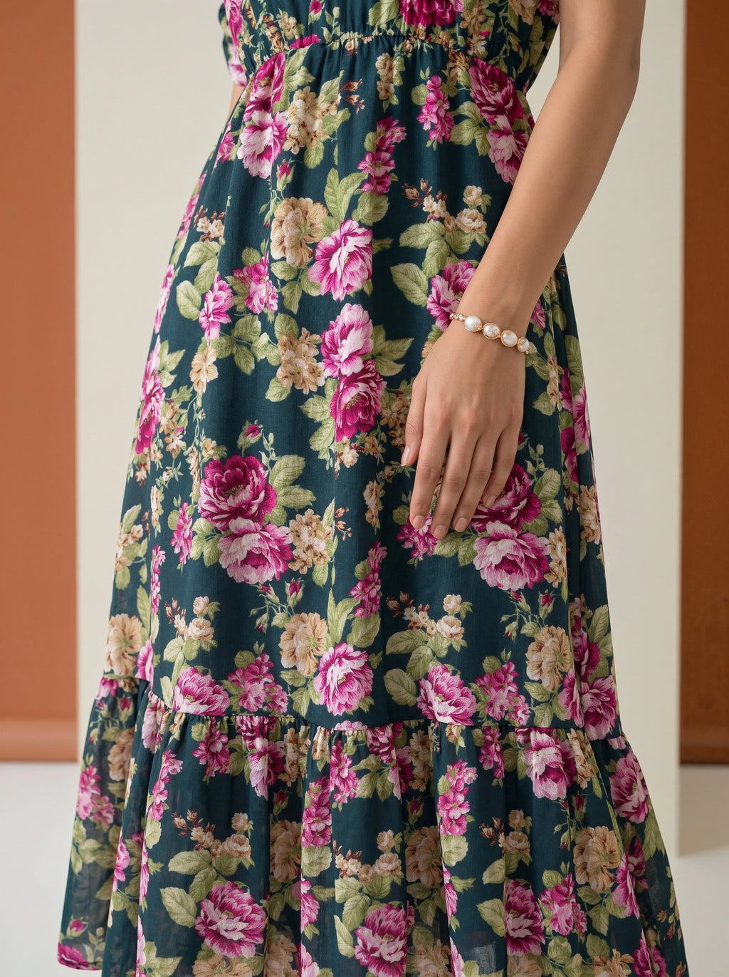 A close-up of a woman wearing a dark teal floral maxi dress with pink and green rose patterns, featuring a smocked bodice and tiered ruffle hem.
