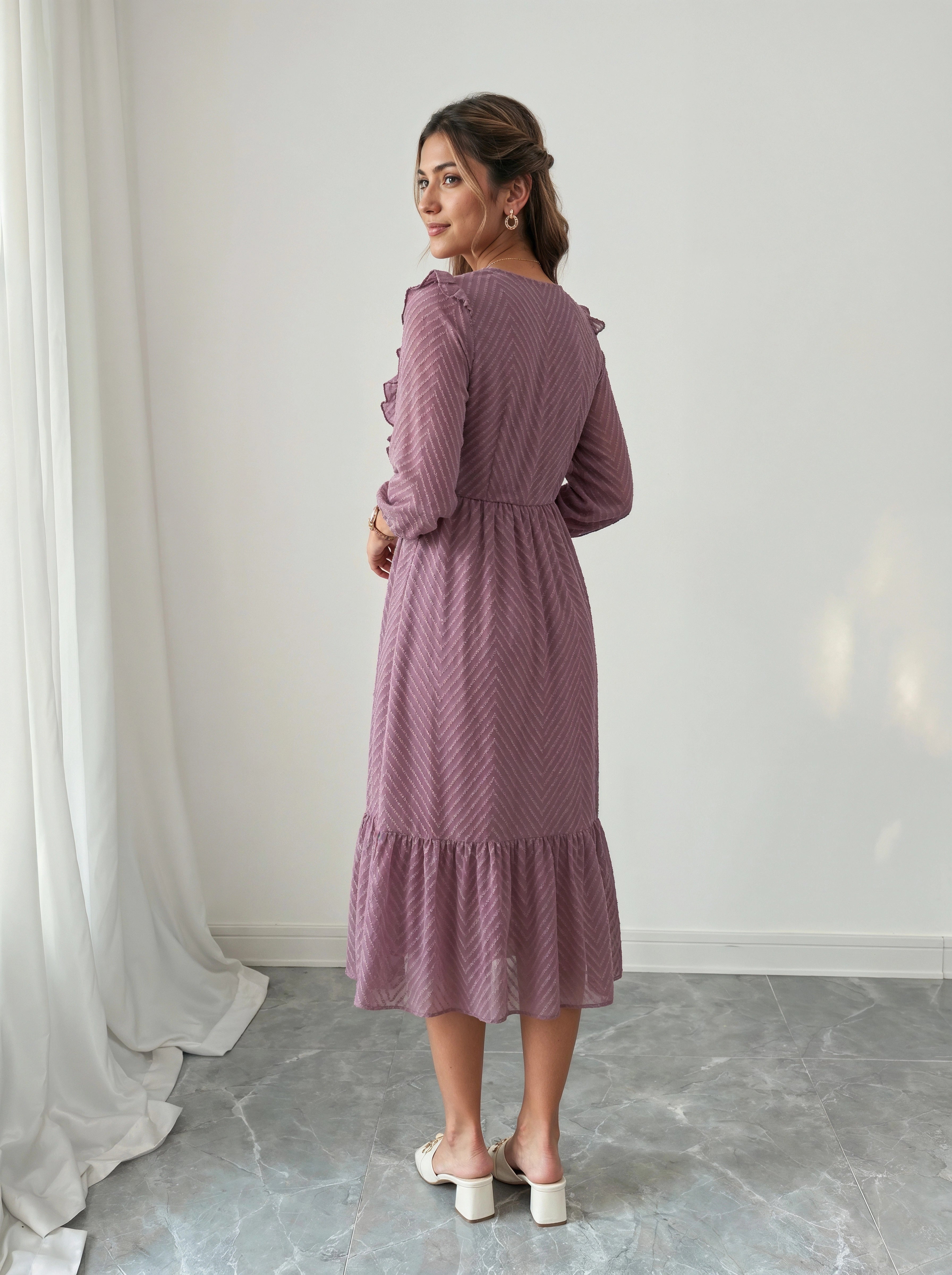 Woman wearing a purple dress standing in a room with white curtains and gray floor.