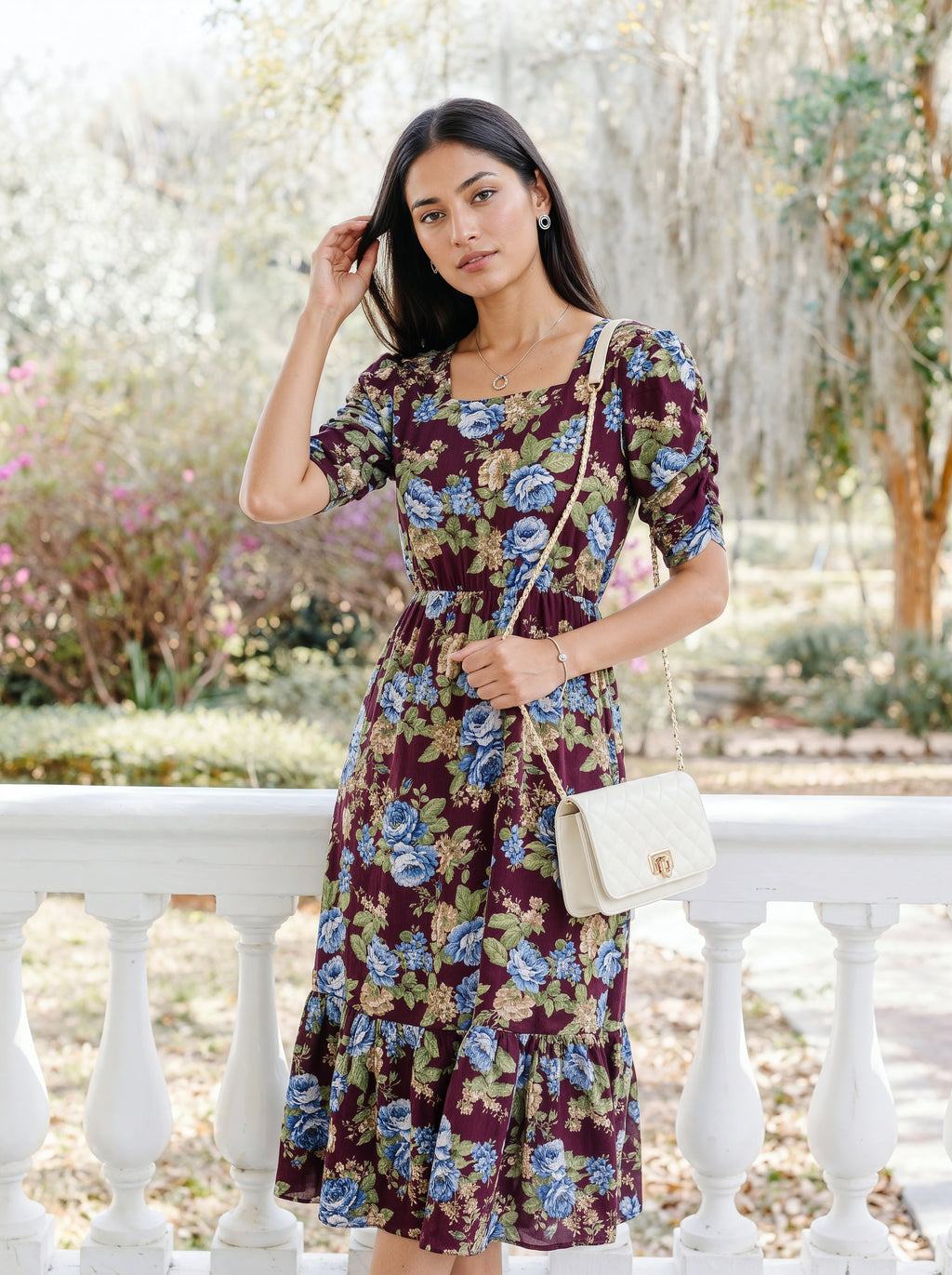 A woman stands outdoors on a white porch railing wearing a short-sleeve, tiered midi dress with a floral print in burgundy, blue, and green. She is carrying a small cream quilted shoulder bag.