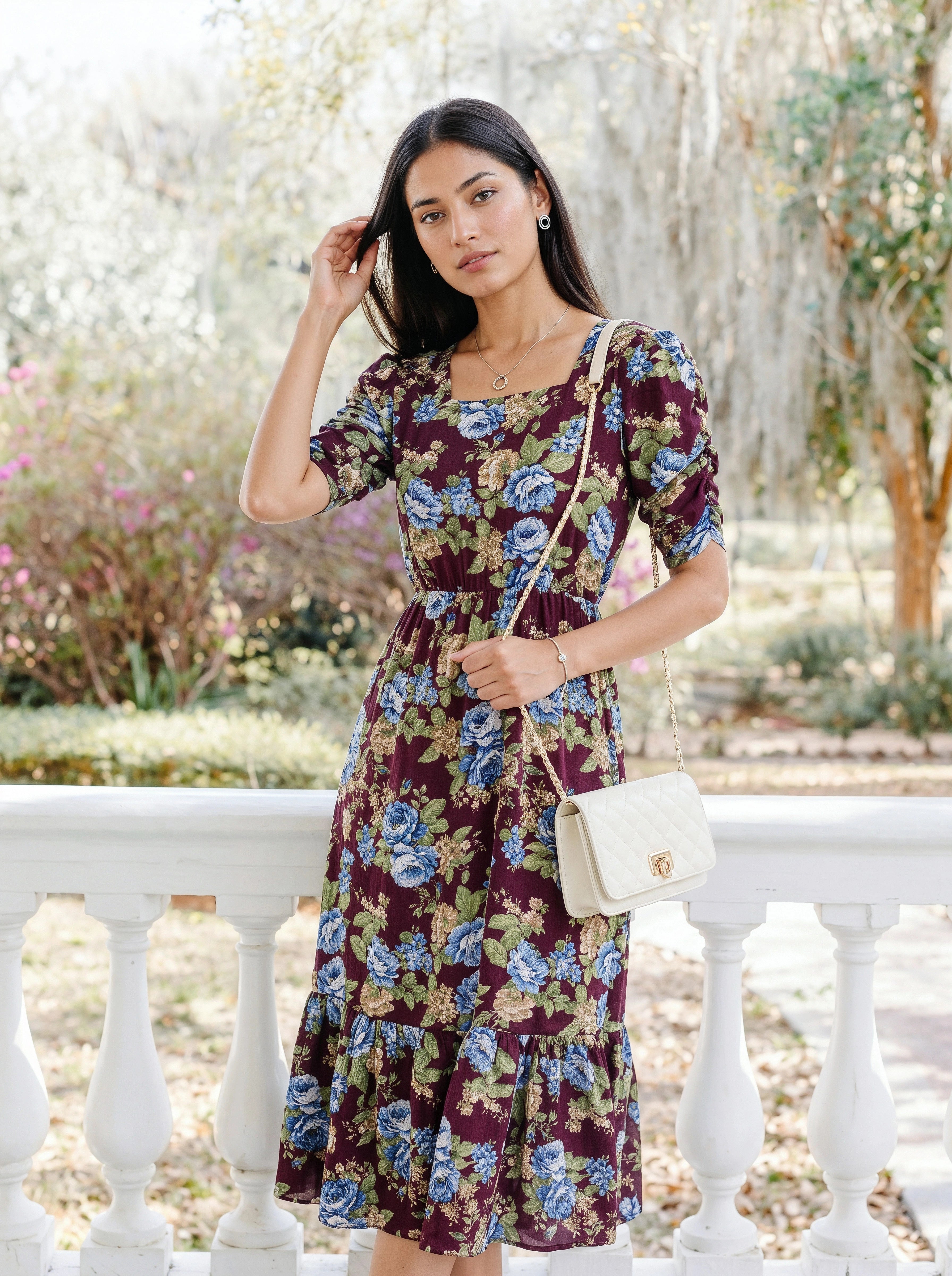 A woman stands outdoors on a white porch railing wearing a short-sleeve, tiered midi dress with a floral print in burgundy, blue, and green. She is carrying a small cream quilted shoulder bag.