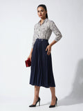 Woman wearing a patterned blouse and navy pleated skirt holding a red clutch against a white background