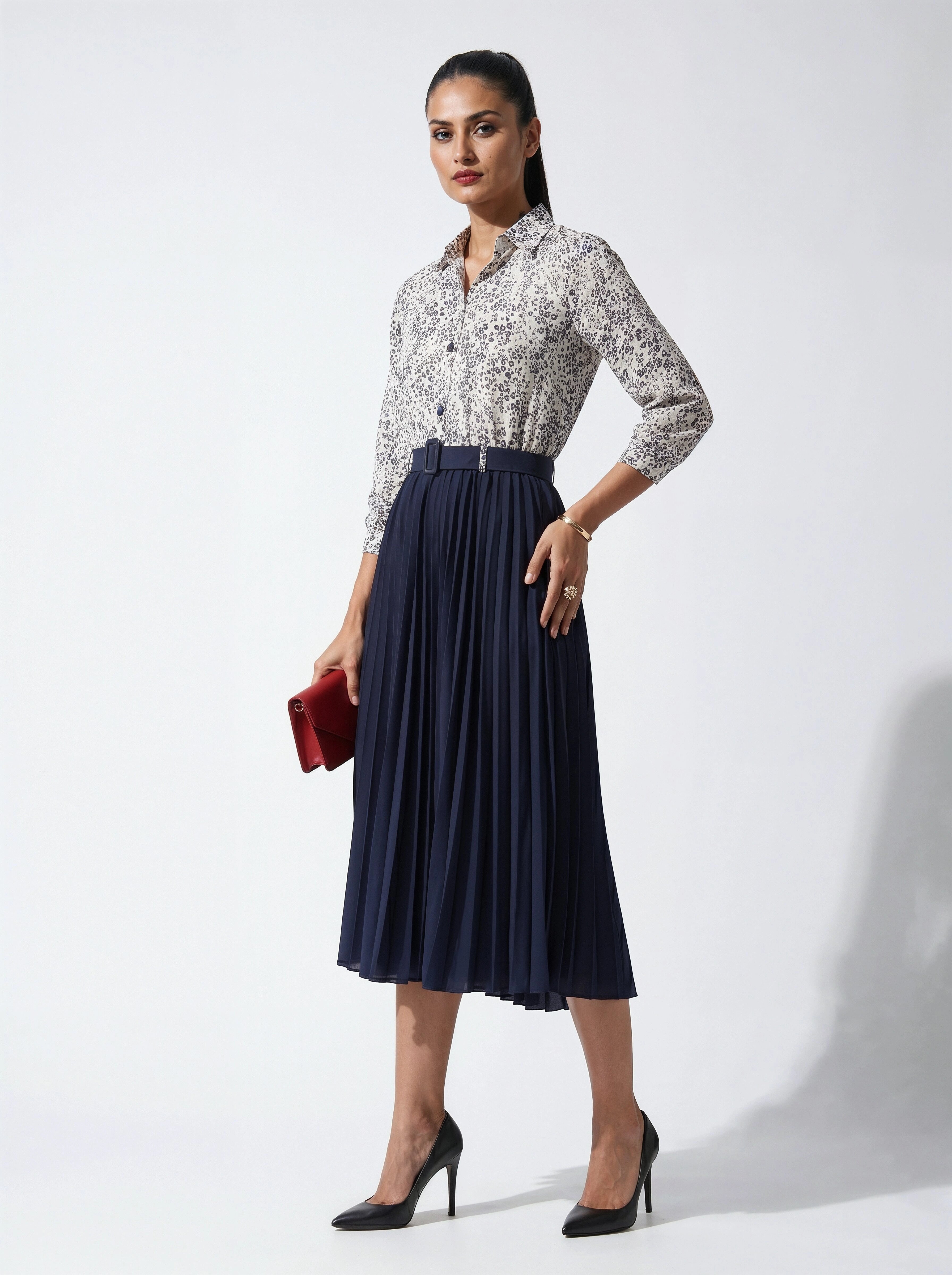 Woman wearing a patterned blouse and navy pleated skirt holding a red clutch against a white background