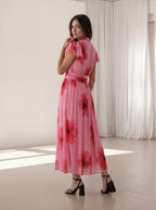 A woman modeling a pink midi dress with a red and black floral print, featuring a high neck, ruffled short sleeves, and a matching waist tie, set in a bright studio with white curtains.