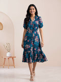 A woman modeling a navy blue tiered midi dress with sheer three-quarter sleeves and a button-front bodice, standing against a warm beige backdrop with draped cream fabric.