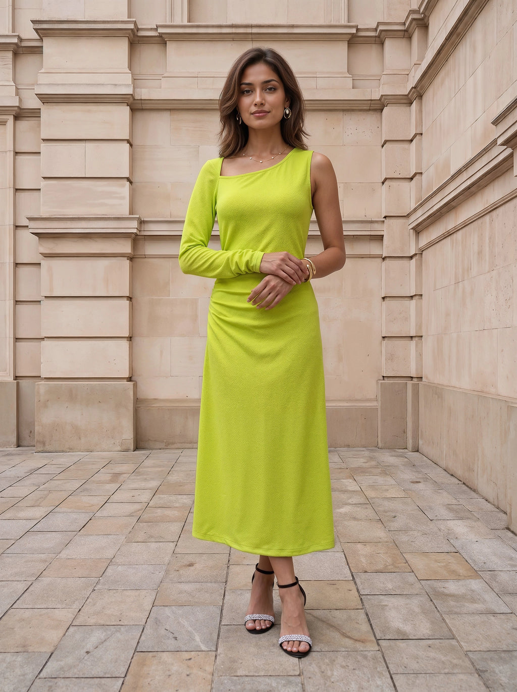 A woman modeling a bright lime green one-shoulder long-sleeve midi dress, standing on a stone tiled floor against a beige stone building background.