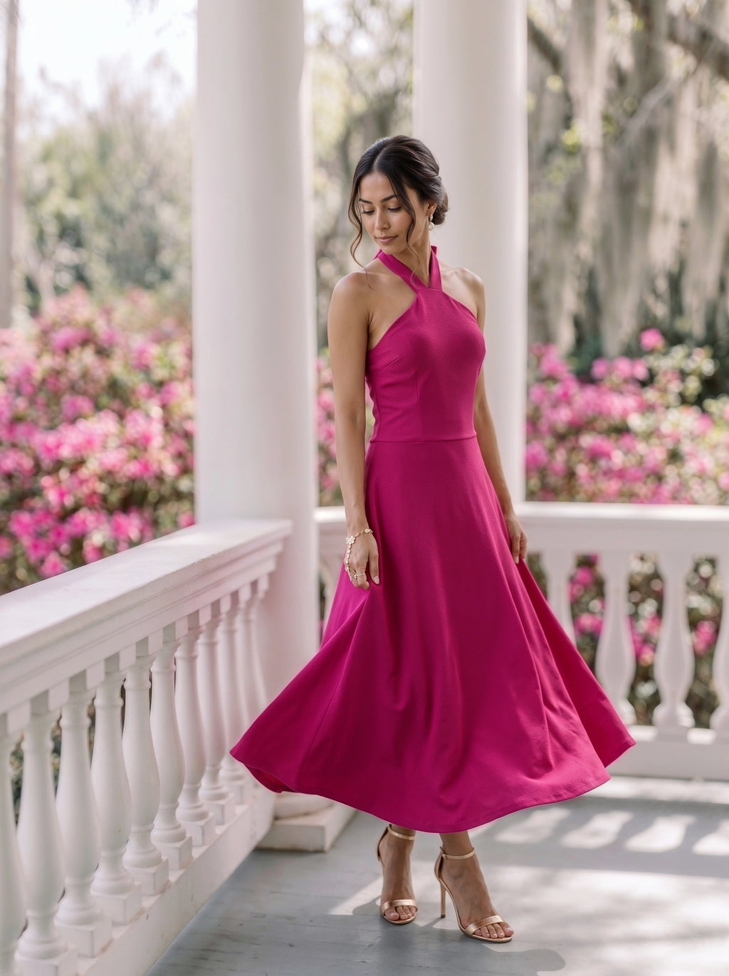 A woman wearing a bright magenta halter-neck midi dress with a flared skirt, standing on a white porch with columns and pink flowers in the background.