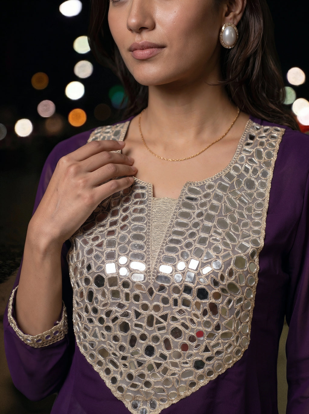 A close-up of a woman wearing a purple kurta with a large beige embroidered yoke decorated with small reflective mirror tiles, set against a dark background with blurred lights.