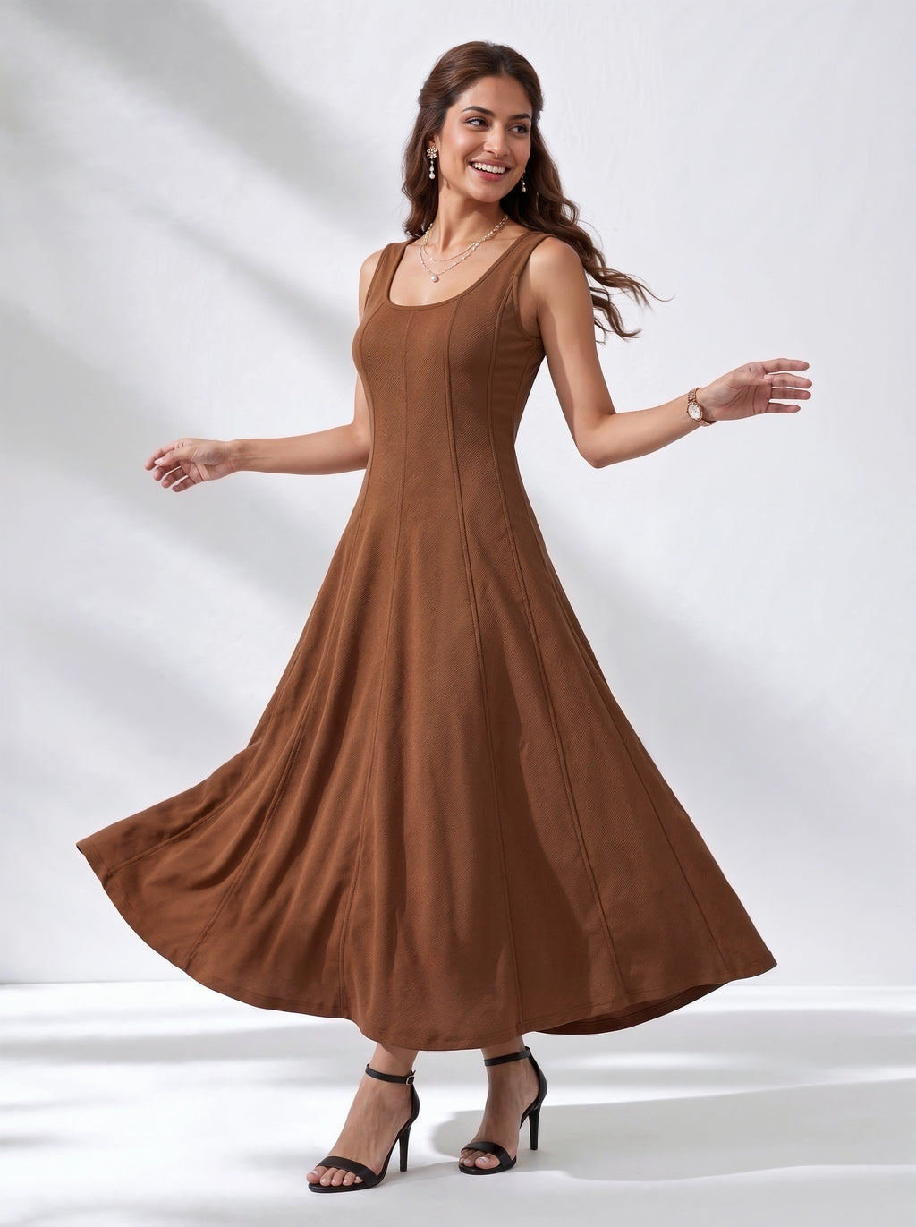 A woman modeling a sleeveless, scoop-neck maxi dress in a warm brown color, captured mid-spin to show the flow of the skirt. She is wearing black high-heeled sandals against a white studio backdrop with soft shadows.