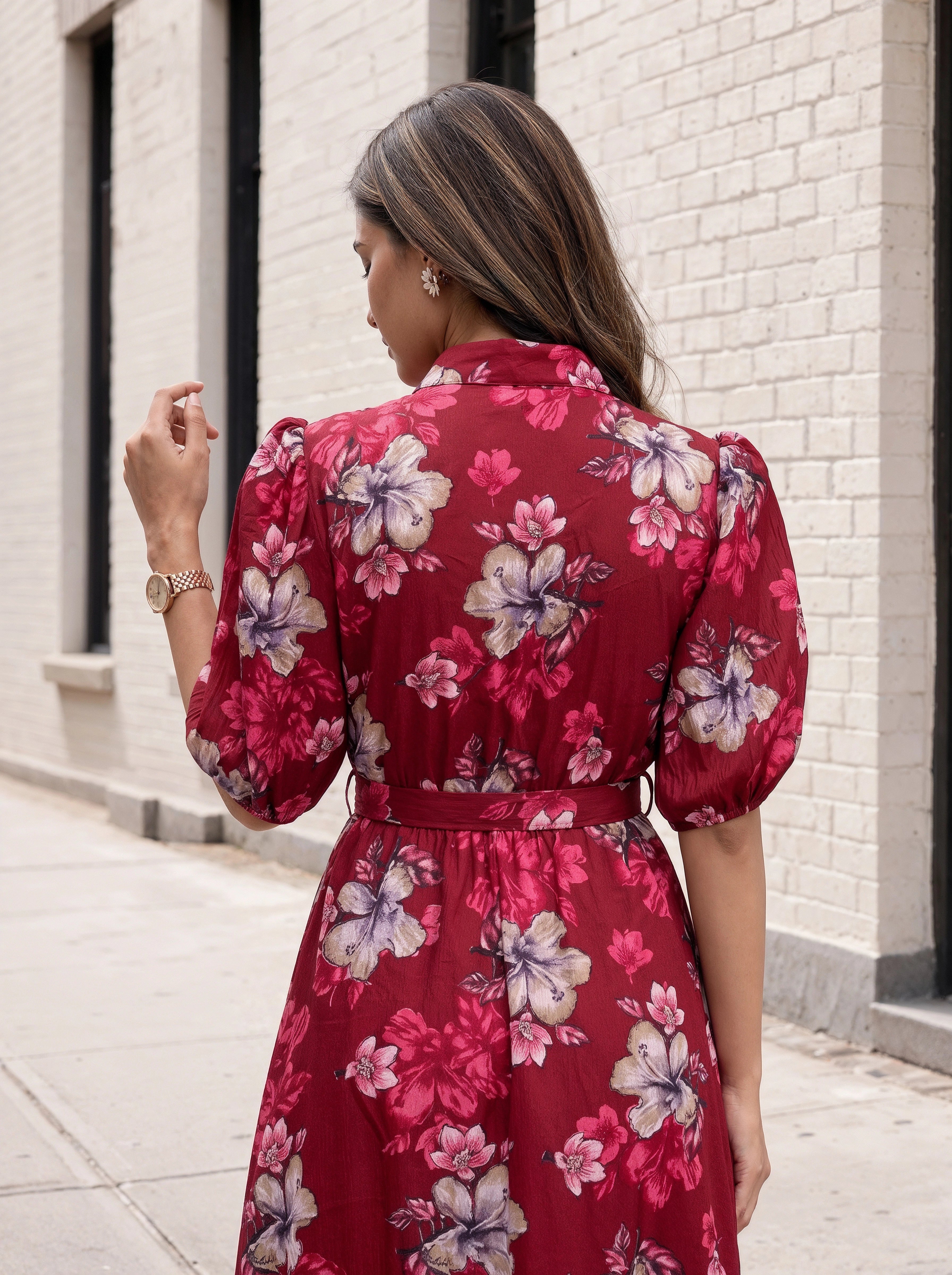 A woman viewed from behind wearing a red floral midi dress with puff sleeves and a matching waist tie, standing on a sidewalk next to a white brick wall.