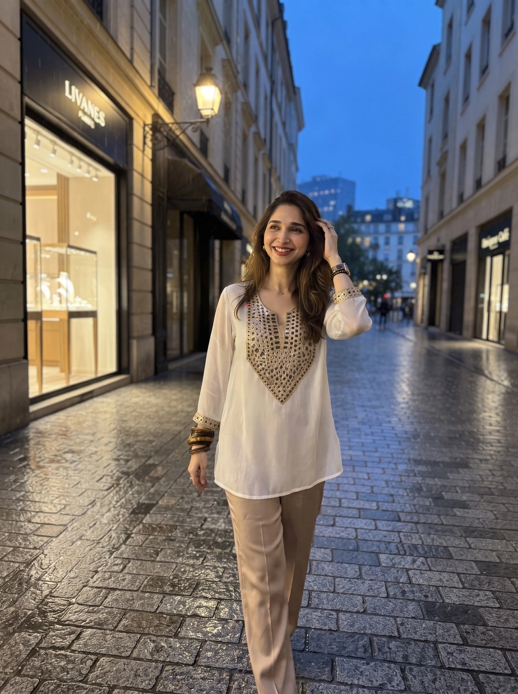 A woman stands on a wet cobblestone street at dusk wearing a white long-sleeve tunic with a gold embroidered V-neck panel and beige trousers. Storefronts line the street, including a visible Louis Vuitton sign on the left.