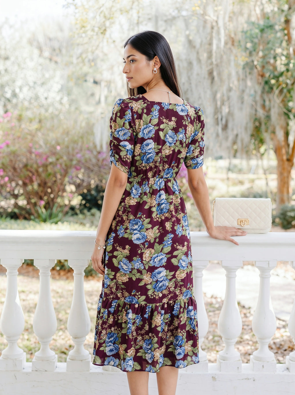 A woman modeling a dark blue midi dress with sheer patterned sleeves and a tiered skirt, standing against a light gray studio background.