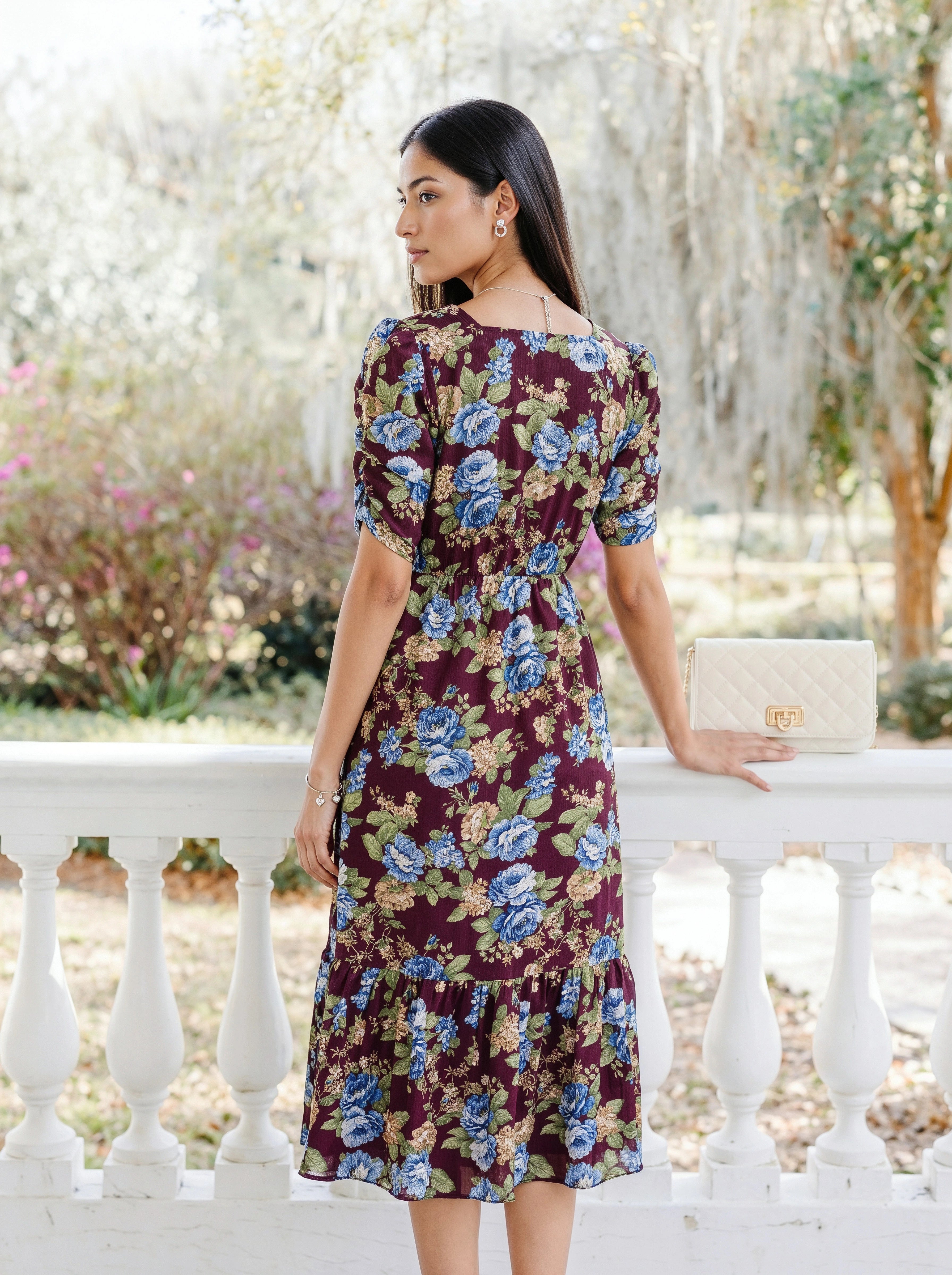 A woman modeling a dark blue midi dress with sheer patterned sleeves and a tiered skirt, standing against a light gray studio background.