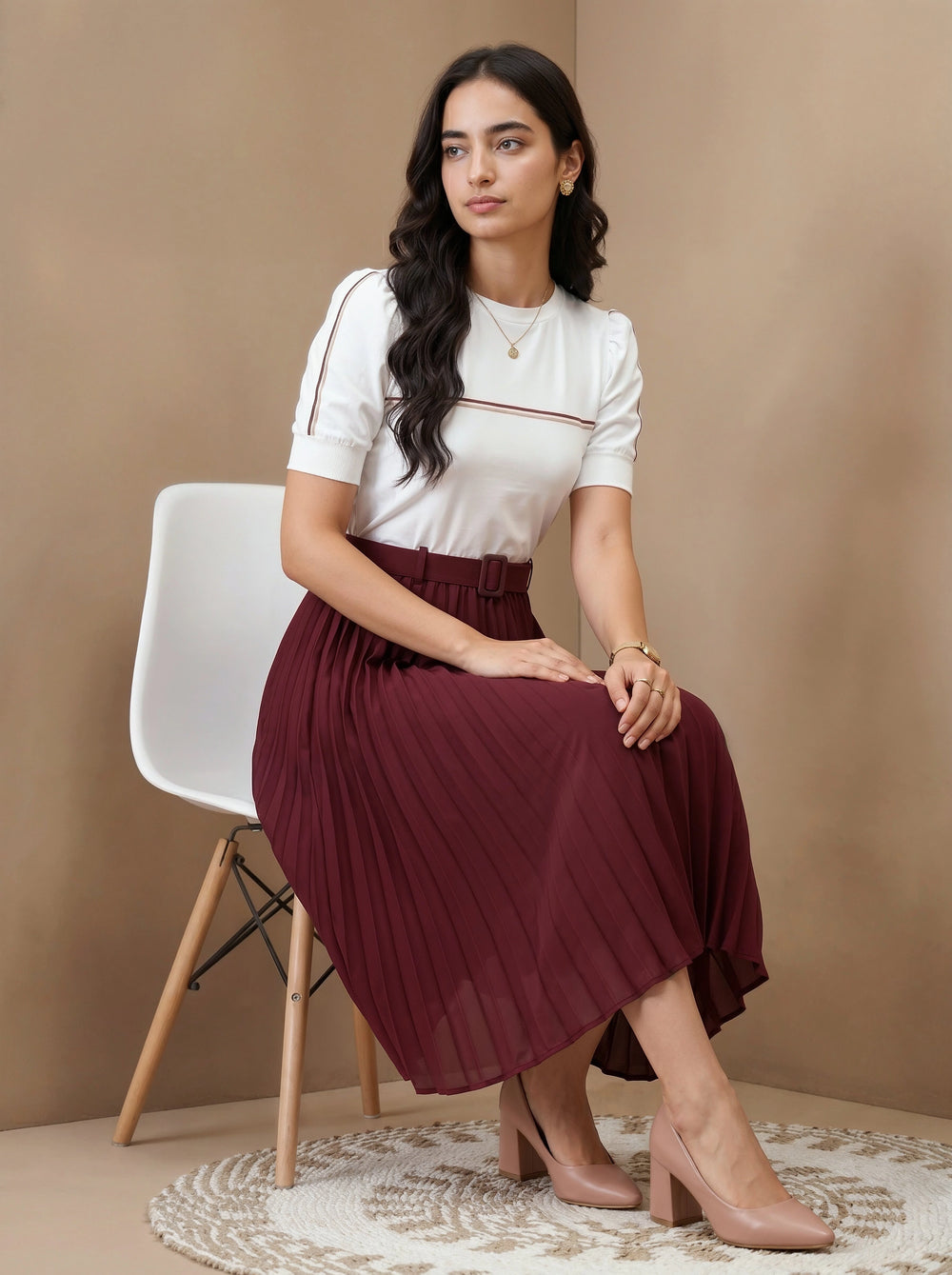 Woman sitting on a chair wearing a white top and burgundy pleated skirt against a beige wall.