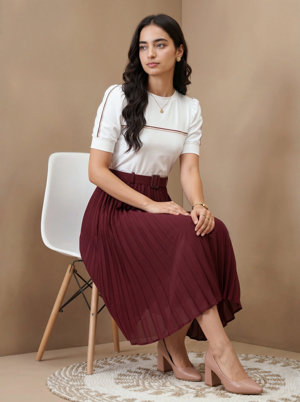 Woman sitting on a chair wearing a white top and burgundy pleated skirt against a beige wall.