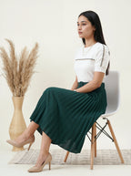 Woman sitting on a chair wearing a white top and green skirt in a minimalistic room.