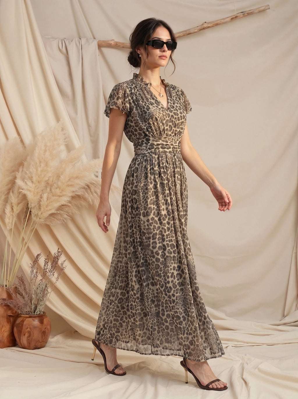 A woman models a leopard-print maxi dress with short flutter sleeves, a V-neckline, and a matching fabric belt. She is wearing black sunglasses and strappy high-heeled sandals, standing in front of a draped beige fabric backdrop with decorative pampas grass.