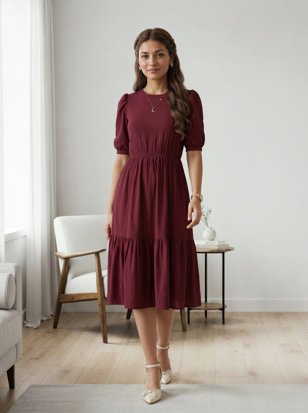 Woman wearing a burgundy dress standing in a room with a white chair and table.