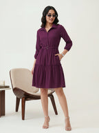 Woman wearing a purple dress standing in a minimalistic room with a chair and table.