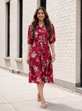 A woman wearing a red floral midi shirt dress with puff sleeves and a tie waist, walking on a sidewalk next to a white brick building.
