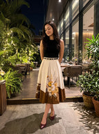 A woman stands outdoors at night in front of a restaurant patio, wearing a sleeveless black top and a pleated midi skirt with a cream upper section, a thin metallic belt, and a brown lower section featuring a floral print. She is wearing burgundy pointed-toe heels.