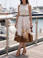A woman stands on a wooden dock by the water wearing a sleeveless white crop top and a high-waisted pleated midi skirt with a brown lower panel and floral print. She is holding sunglasses and wearing brown lace-up block-heel sandals.