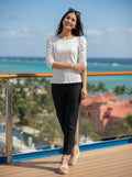 Woman standing on a deck with ocean view wearing a white top and black pants.