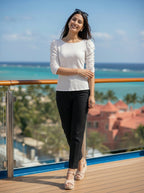 Woman standing on a deck with ocean view wearing a white top and black pants.
