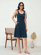 Woman wearing a navy blue dress standing in a minimalistic room with a chair and vase.