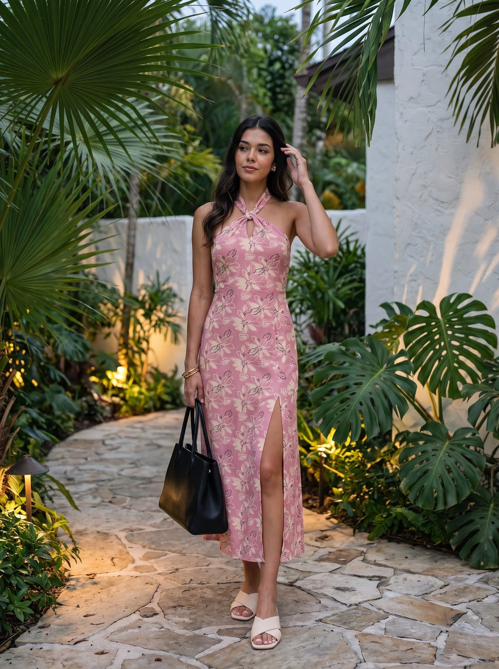 A woman stands on a stone pathway surrounded by tropical greenery, wearing a pink floral halter-neck midi dress with a thigh-high slit. She is holding a black tote bag and wearing beige slide sandals.