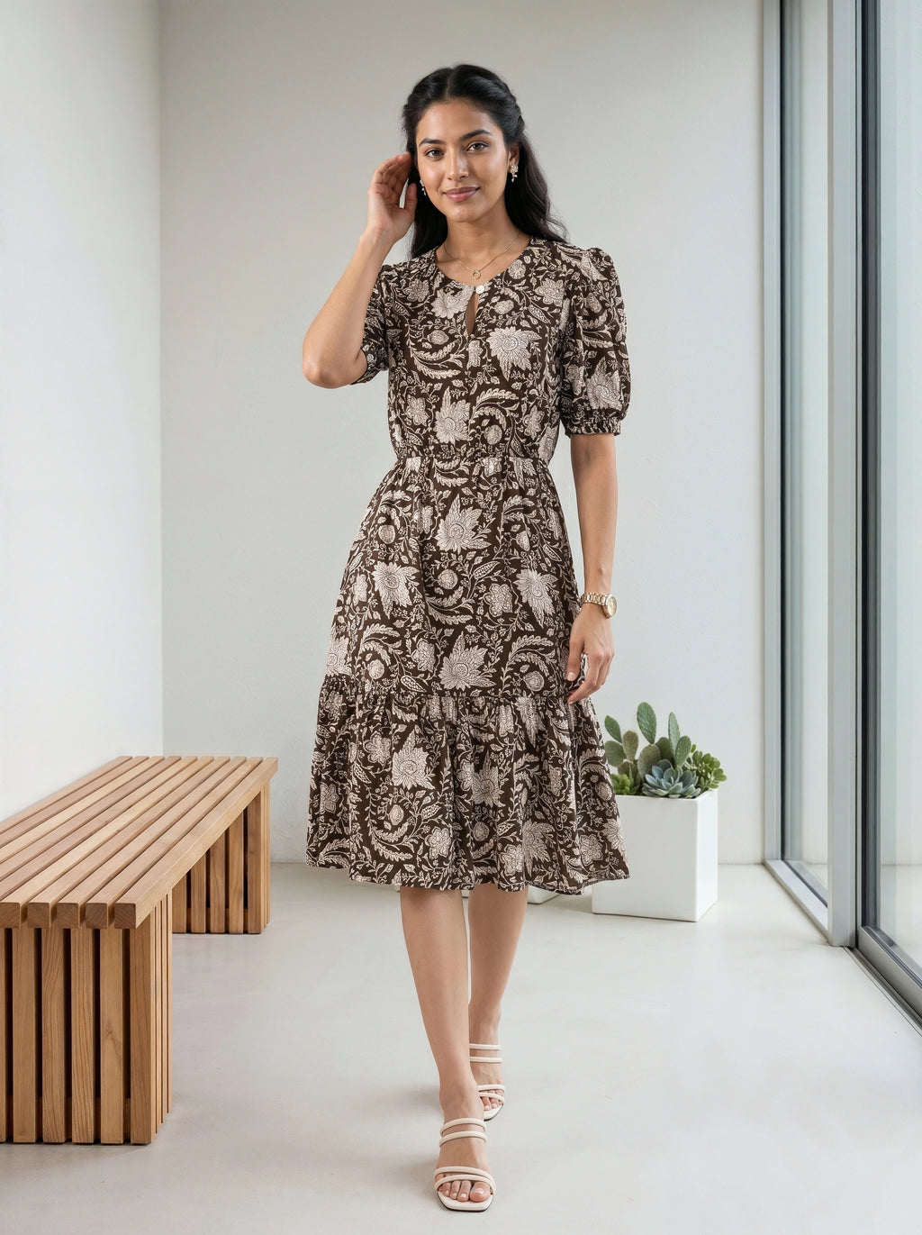 A woman models a brown and beige floral print midi dress with puff sleeves and a keyhole neckline, standing in a bright, modern indoor space with a wooden bench and large windows.