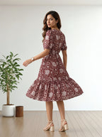 A woman modeling a short-sleeve, knee-length dress with a ruffled hem, featuring a dusty rose and white floral paisley pattern. She is standing in a bright room with light wood floors and a potted plant on the left.