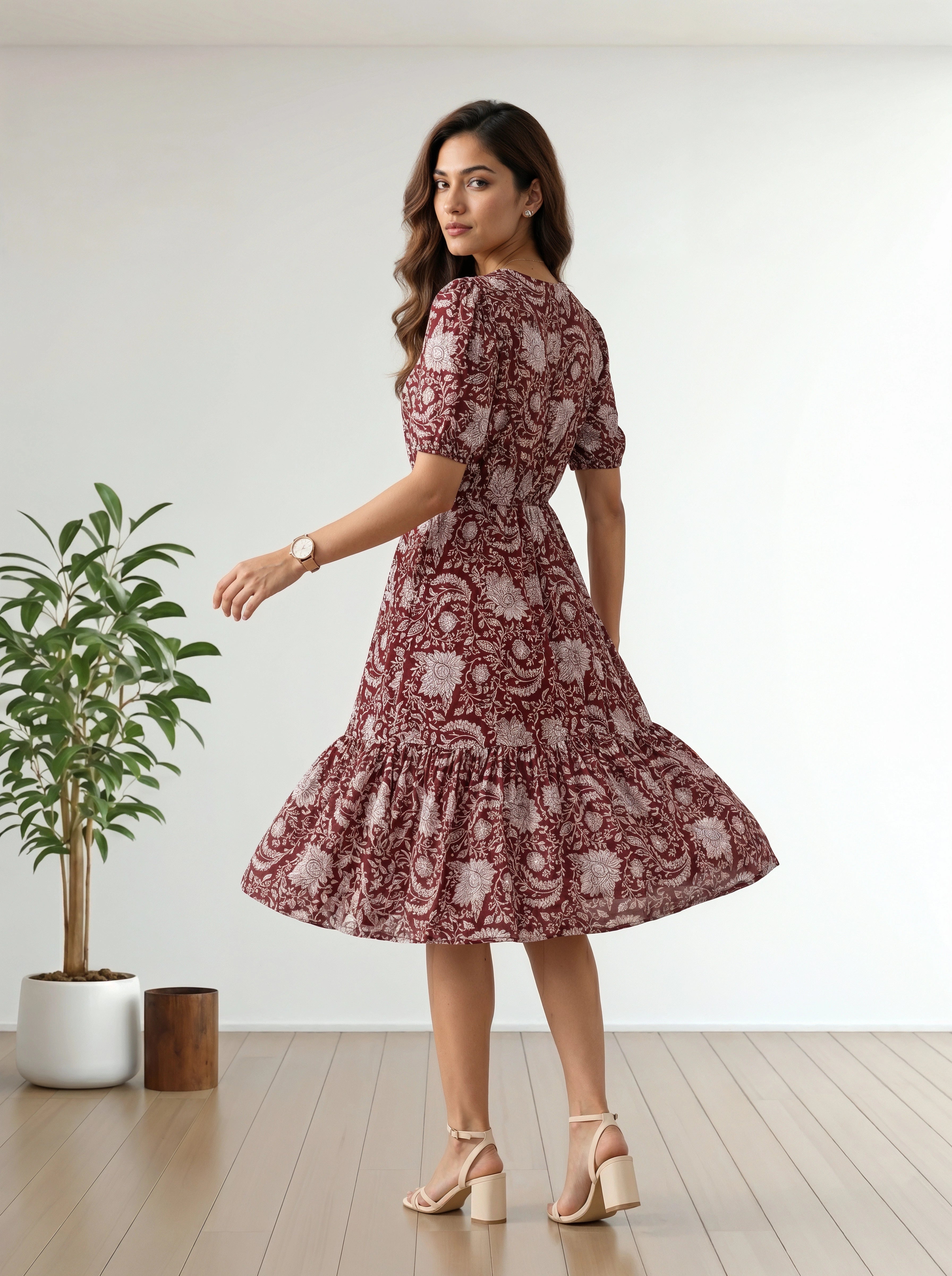 A woman modeling a short-sleeve, knee-length dress with a ruffled hem, featuring a dusty rose and white floral paisley pattern. She is standing in a bright room with light wood floors and a potted plant on the left.