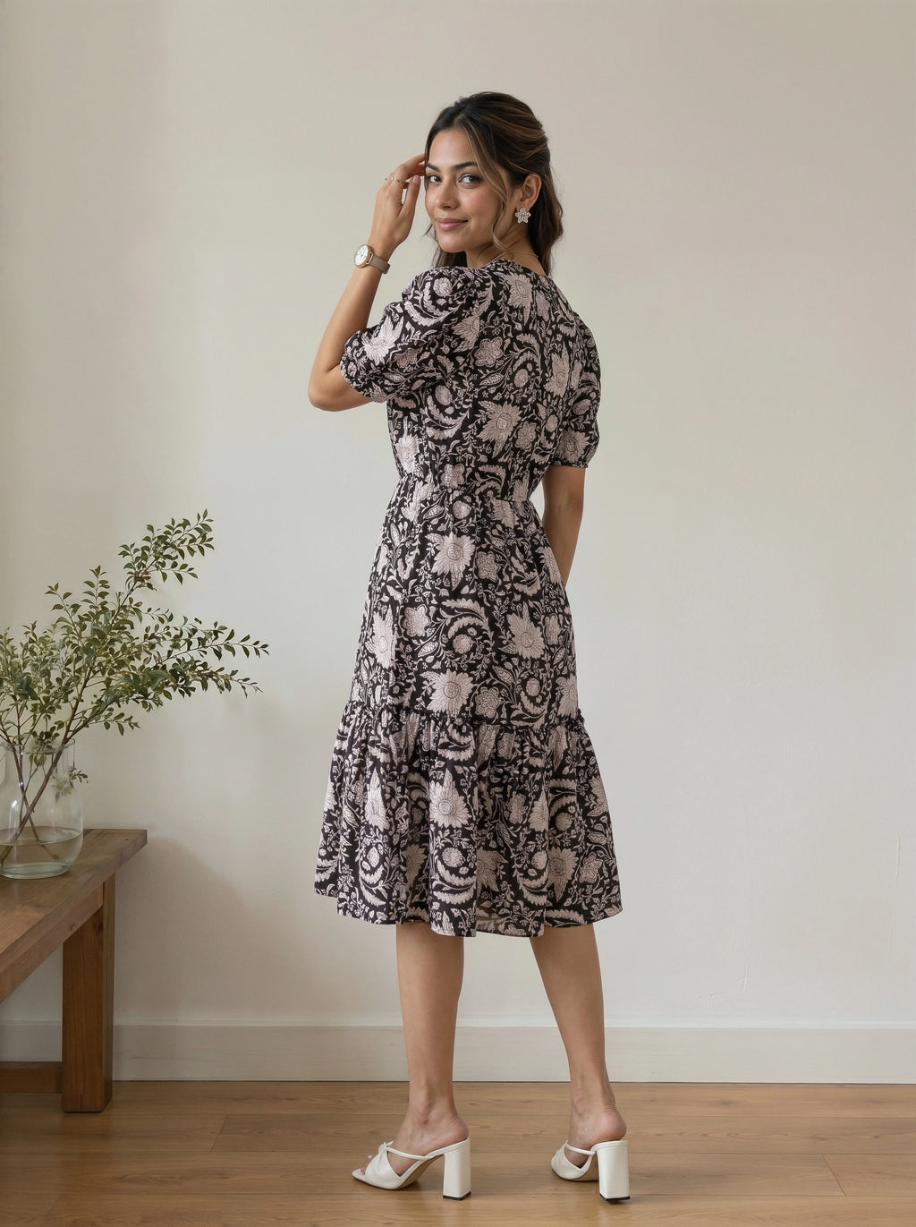 A woman modeling a short-sleeve midi dress with a black and beige floral pattern, standing in a minimalist room with a wooden floor and a small plant on a side table.