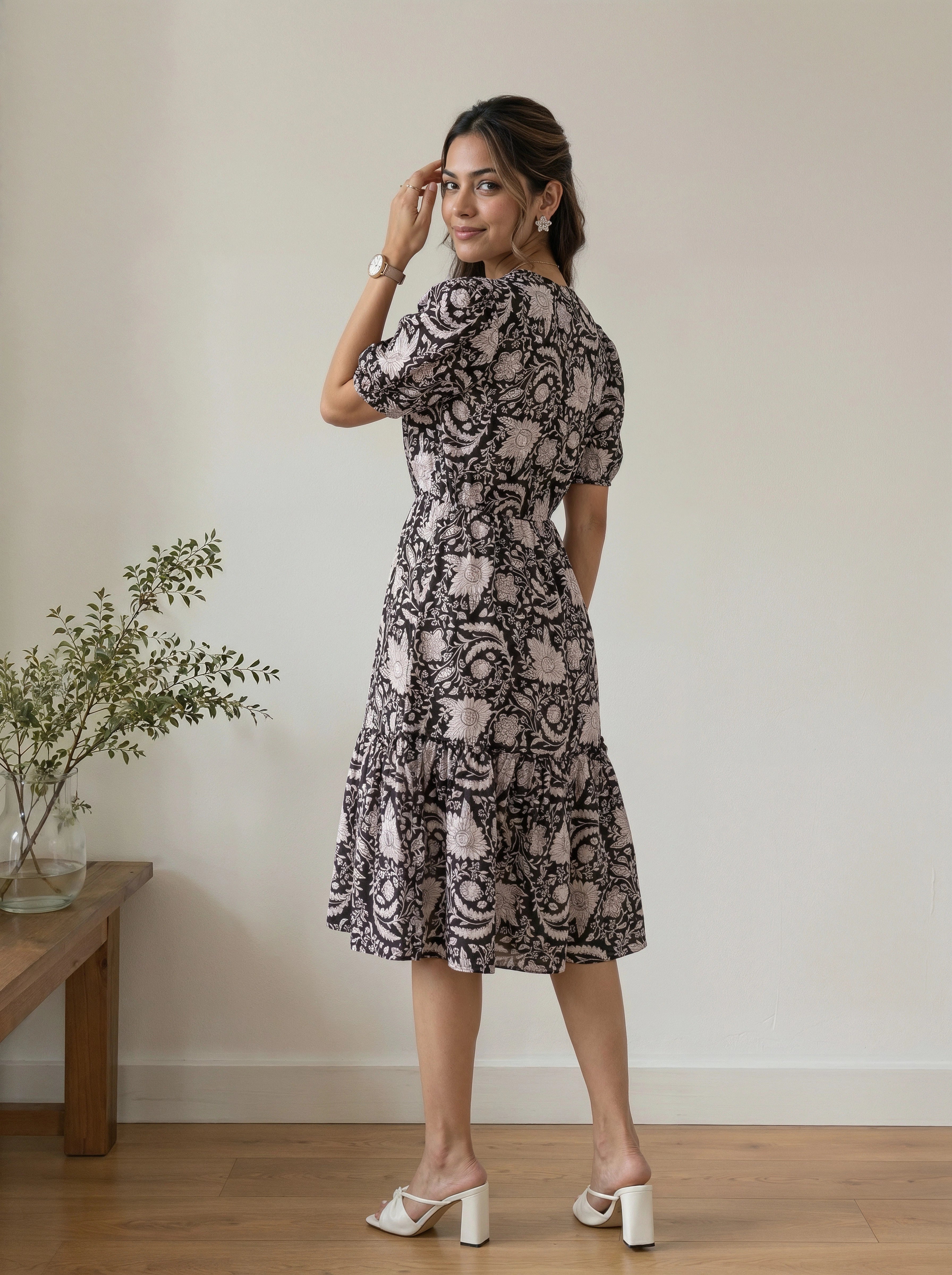 A woman modeling a short-sleeve midi dress with a black and beige floral pattern, standing in a minimalist room with a wooden floor and a small plant on a side table.