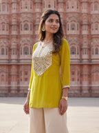 A woman wearing a bright yellow kurta with intricate silver mirror work embroidery on the chest and cuffs, paired with beige wide-leg trousers, standing in front of the iconic pink sandstone walls of Amber Palace.