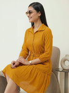 Woman in a mustard yellow dress sitting on a chair with a neutral background