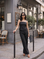A woman wearing a black sleeveless top and matching wide-leg trousers stands on a city sidewalk in front of a gray storefront, holding a white handbag and wearing nude pointed-toe heels.