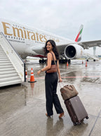 A woman stands on a wet airport tarmac in front of an Emirates airplane, pulling a brown rolling suitcase with a matching brown duffel bag. She is wearing a black strappy top and black wide-leg trousers.