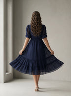 Woman wearing a blue dress standing in a room with white walls and a window.