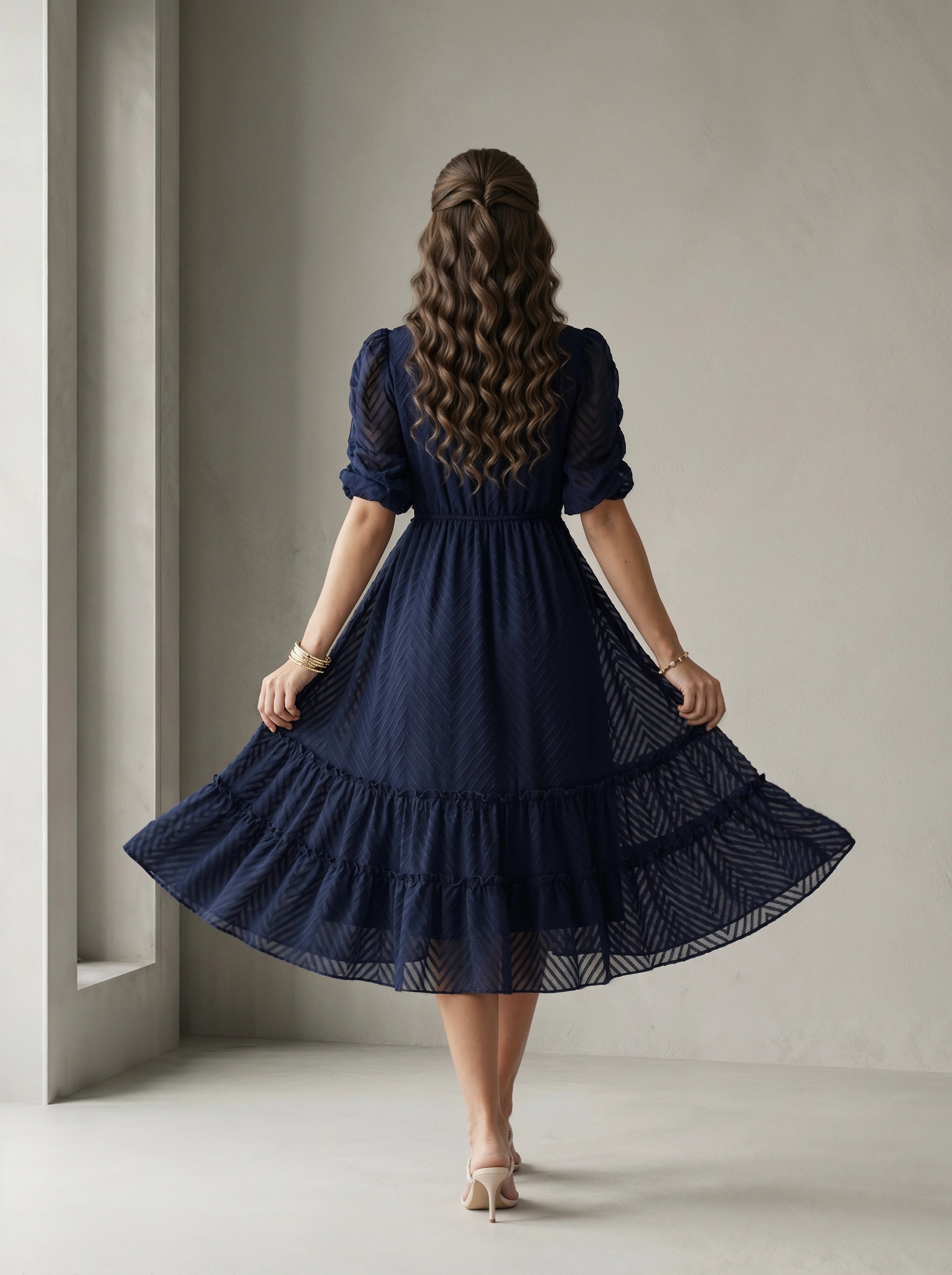 Woman wearing a blue dress standing in a room with white walls and a window.