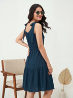 Woman wearing a blue dress with sunglasses, standing in a room with a chair and decorative plant.