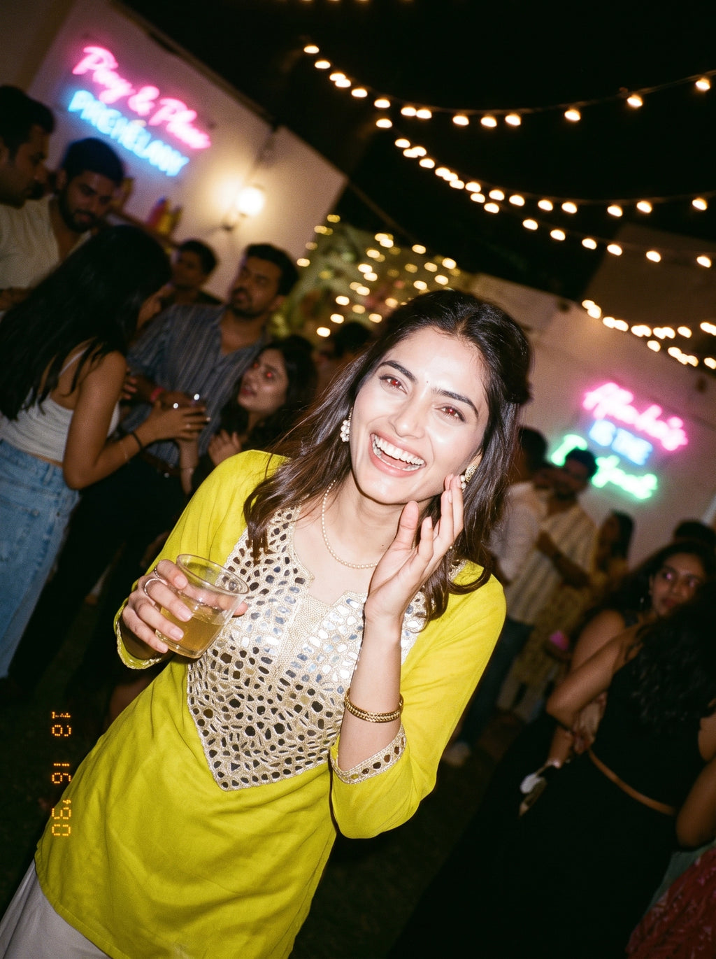 A woman smiling and holding a drink at a nighttime social event, wearing a bright yellow kurta with a silver beaded embroidered yoke. The background is dark with warm string lights and blurred people.