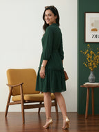 Woman in a green dress standing in a room with a yellow armchair and a side table.