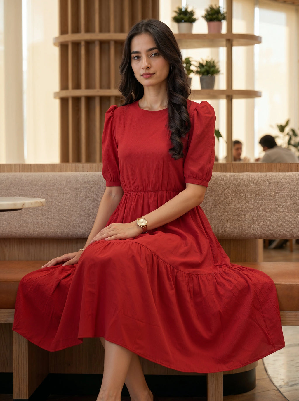 Woman sitting in a modern, sunlit interior wearing an elegant red tiered midi dress with puff sleeves.