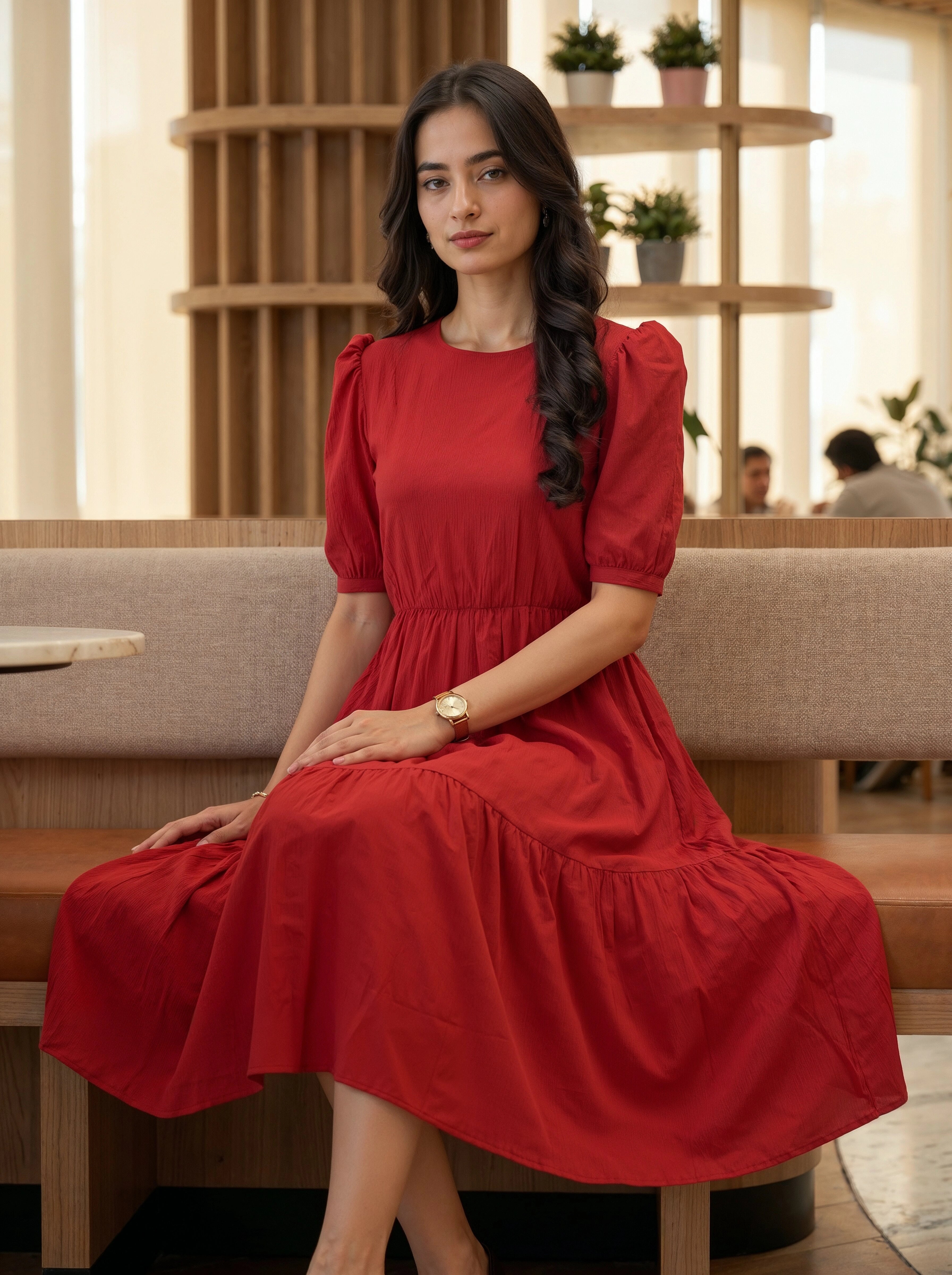 Woman sitting in a modern, sunlit interior wearing an elegant red tiered midi dress with puff sleeves.