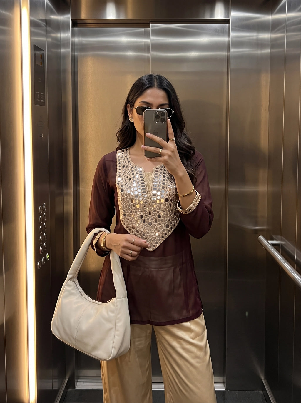 A woman takes a mirror selfie in an elevator wearing a maroon long-sleeve top with a heavily embellished silver sequin V-neck panel, paired with beige trousers and carrying a large off-white shoulder bag.