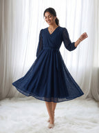 Woman wearing a navy blue dress with sheer sleeves against a white curtain background